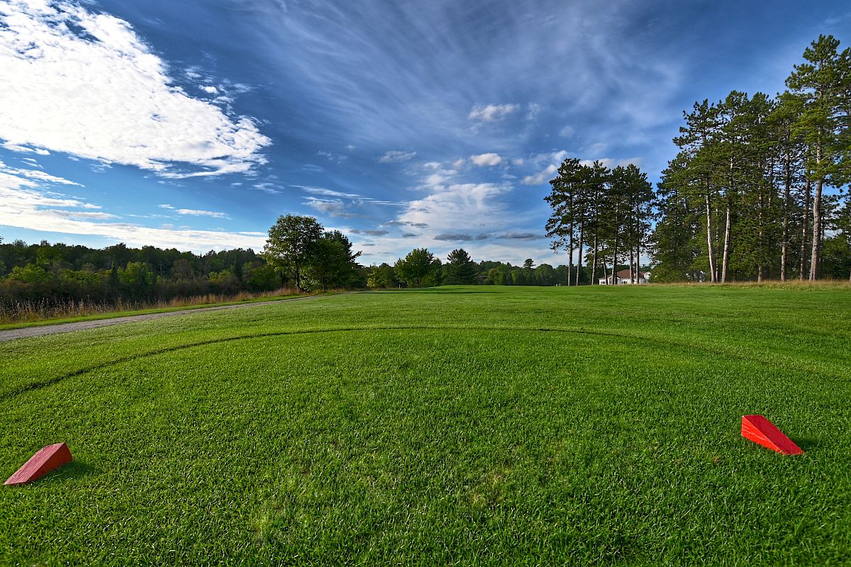 A green golf course with two red markers on the grass, surrounded by trees and under a partly cloudy sky with patches of blue.