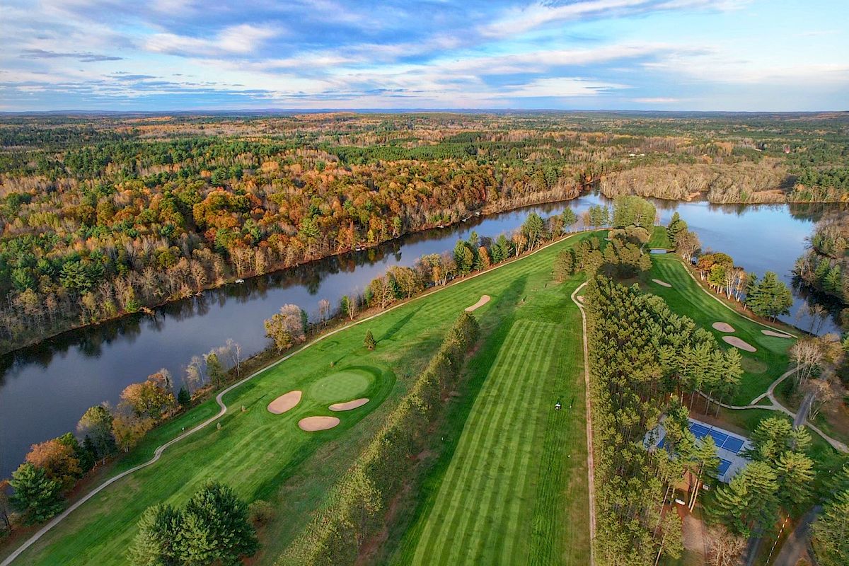 An aerial view of a golf course surrounded by trees, with a river running alongside and a blue sky with scattered clouds.