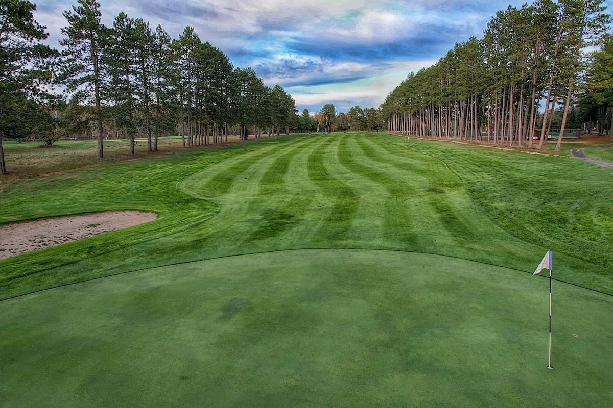 This image shows a well-manicured golf course with striped fairways, a putting green with a flag, and trees lining both sides of the fairway.