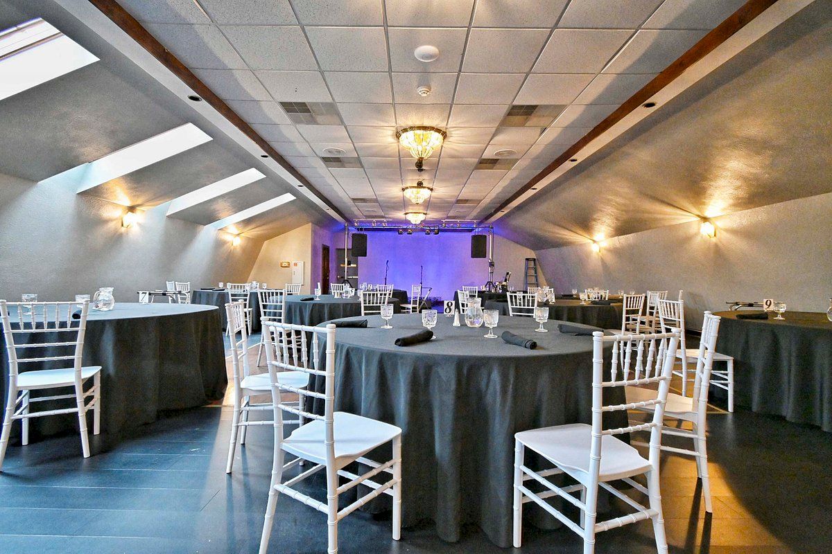 The image shows a decorated event hall with round tables covered in gray tablecloths and white chairs under soft lighting.