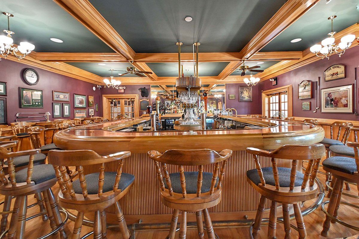 A cozy bar with wooden stools, a curved counter, ceiling fans, and chandeliers. Decor includes clocks, framed pictures, and a warm ambiance.