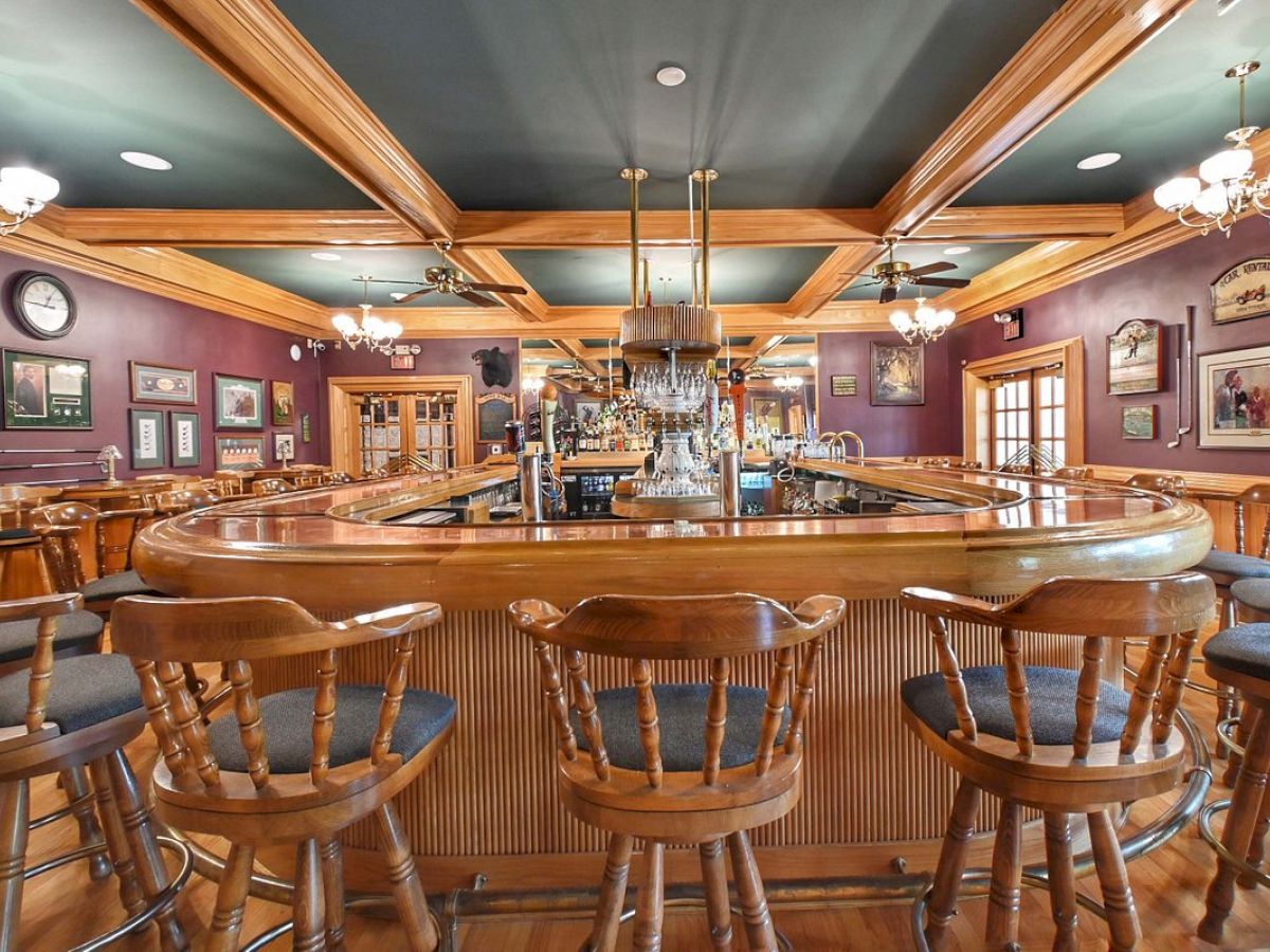 A cozy bar with wooden stools, a curved counter, ceiling fans, and chandeliers. Decor includes clocks, framed pictures, and a warm ambiance.