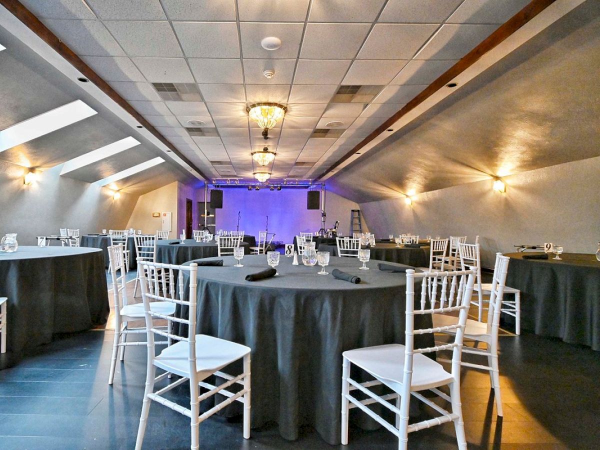 The image shows an elegant, dimly lit banquet hall with round tables covered in dark tablecloths and surrounded by white chairs, ready for an event or gathering.
