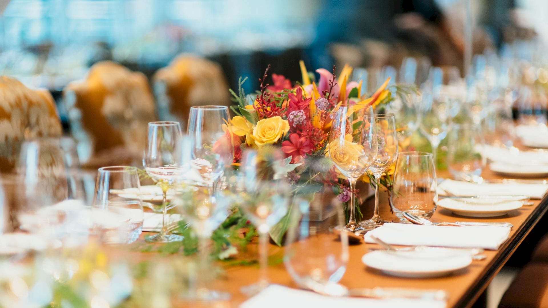 A long dining table is elegantly set with glassware, plates, and a colorful floral centerpiece, ready for a formal event or gathering.