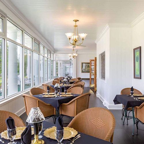 A dining area with wicker chairs, black tablecloths, and elegant table settings, featuring large windows and decorative lighting fixtures.