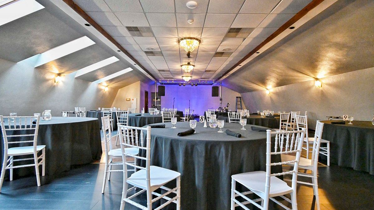 A well-lit banquet hall with round tables covered in dark tablecloths and surrounded by white chairs, prepared for an event with stage setup.