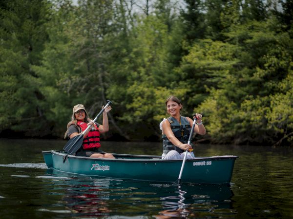 Two people are canoeing on a calm lake surrounded by lush greenery.