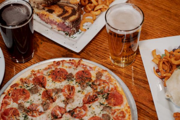 The image shows a pepperoni pizza, a sandwich with curly fries, a wrap, and two glasses of beer on a wooden table, emphasizing a casual meal.