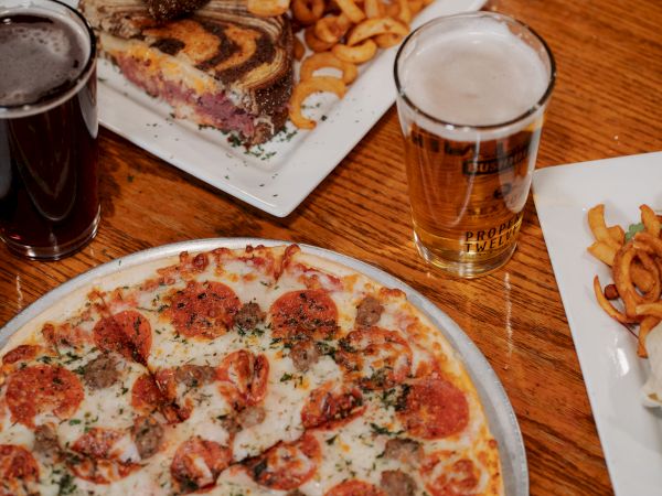 The image shows a pepperoni pizza, a sandwich with curly fries, a wrap, and two glasses of beer on a wooden table, emphasizing a casual meal.