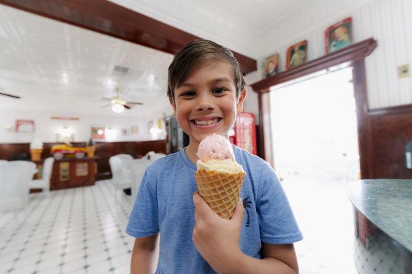 A smiling boy is standing in a bright ice cream shop, holding an ice cream cone with two scoops of ice cream.