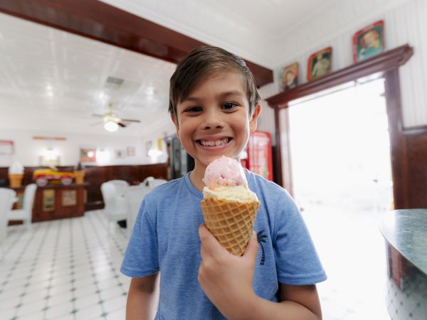 A smiling boy is standing in a bright ice cream shop, holding an ice cream cone with two scoops of ice cream.