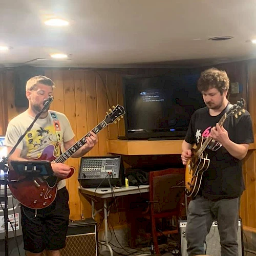 Two people are playing electric guitars in a cozy indoor setting with wood-paneled walls, and various music equipment around them.