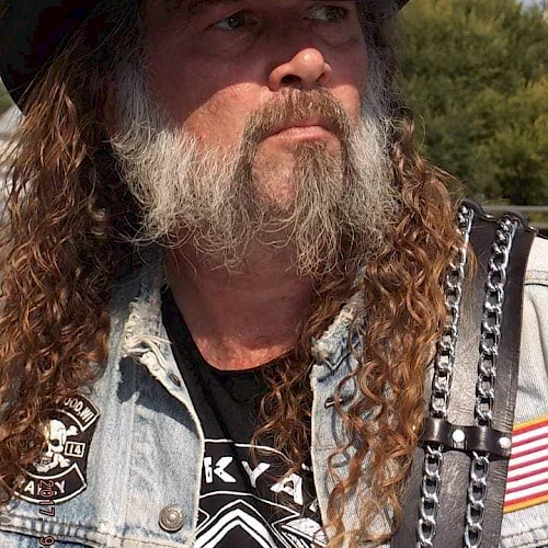 A bearded man wearing a cowboy hat, denim vest, and leather strap with metal details, featuring a patch with a skull design and an American flag patch.