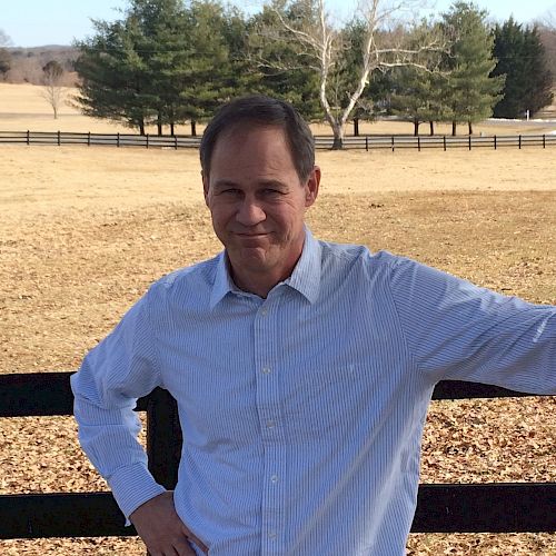 A man stands by a wooden fence in a rural area, with open fields and trees in the background.