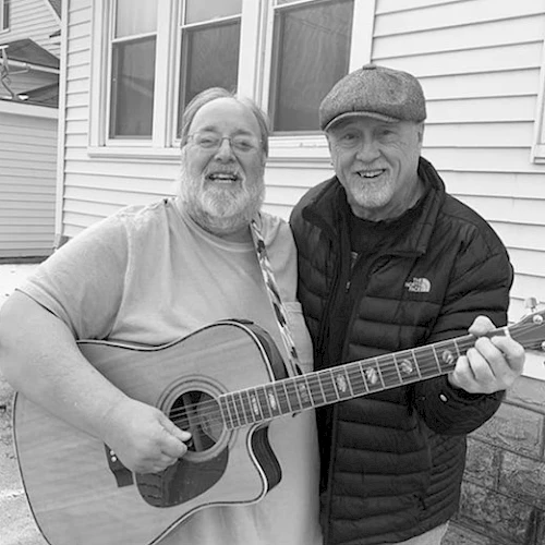 Two smiling men, one playing an acoustic guitar, posing together outside a house. One is dressed in a jacket and cap, the other in a t-shirt.