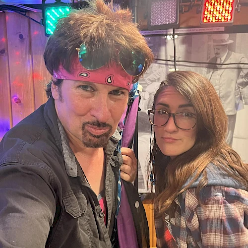 A man and woman are posing for the camera in what seems like a casual indoor setting with colorful lights in the background.