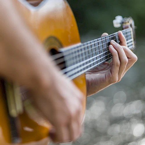 A person is playing an acoustic guitar outdoors near a body of water, with a blurred background.