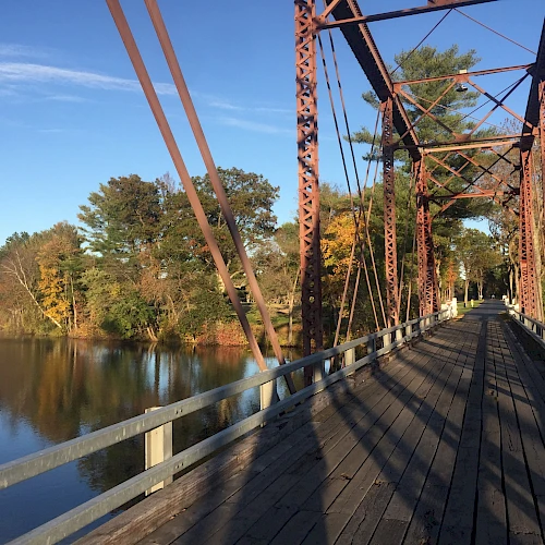 A picturesque wooden bridge with rusted metal railings extends over a calm river surrounded by trees, bathed in sunlight under a clear blue sky.