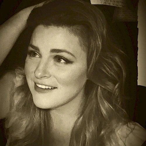 A sepia-toned image of a smiling woman with wavy hair, resting her hand on her head. Text blurred in the background.