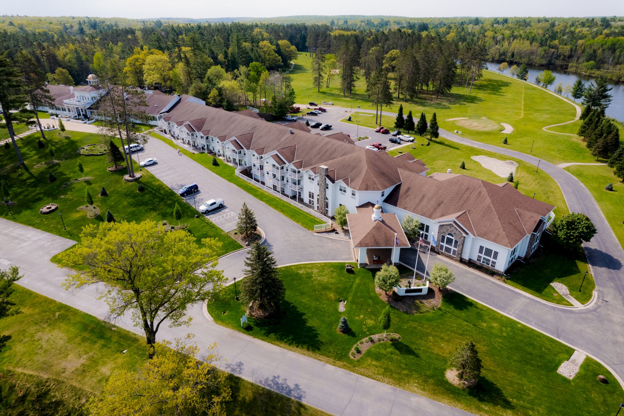 An aerial view of a large residential complex surrounded by greenery, trees, a parking area, and a nearby body of water.