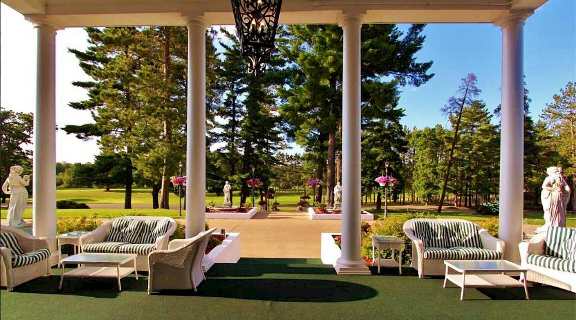 An outdoor garden terrace with white columns, striped lounge chairs, statues, and blooming flowers under a clear blue sky.