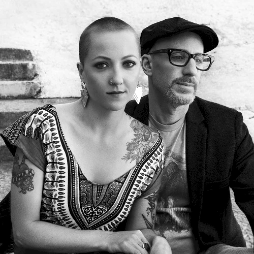 A black and white photo of two people: a woman with a shaved head and tattoos, and a man wearing glasses and a cap, both seated on steps.