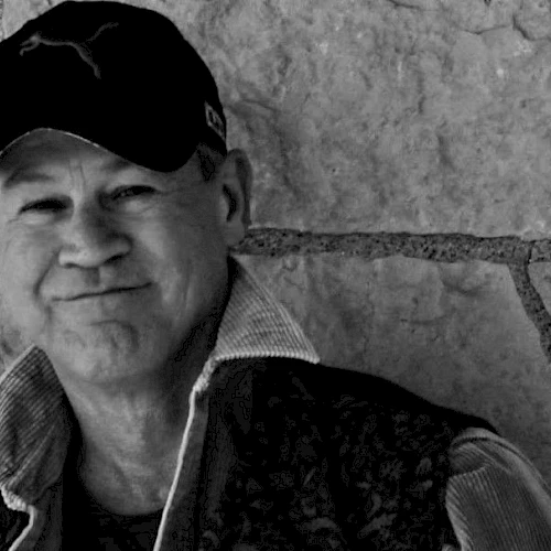 A person wearing a baseball cap and a jacket stands in front of a stone wall, smiling at the camera.