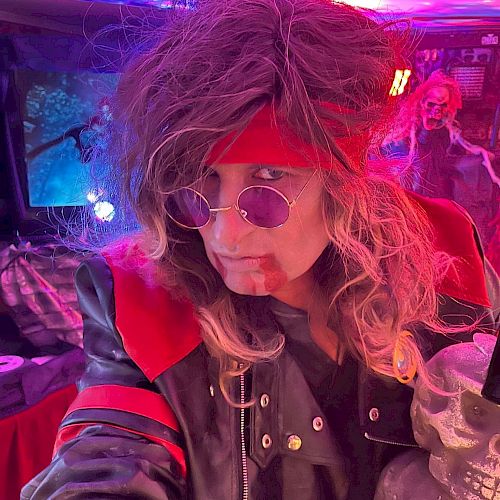 A person in a rocker costume with a red headband, purple glasses, and wild hair in a colorful room, holding a skull decoration.