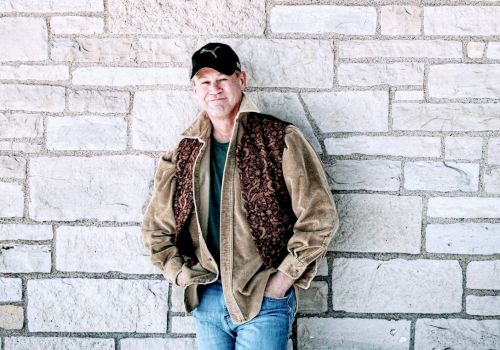 A man in casual attire, with a brown jacket and blue jeans, leans against a stone wall with his hands in his pockets, wearing a cap.