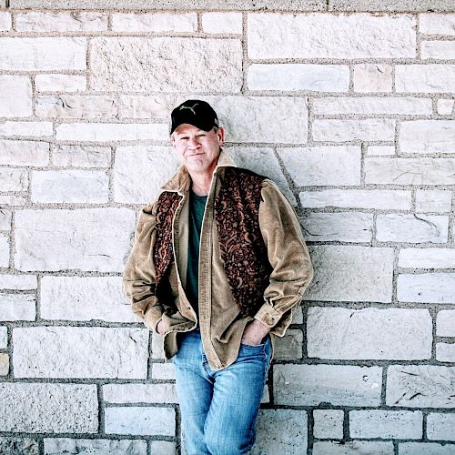 A man in casual attire, with a brown jacket and blue jeans, leans against a stone wall with his hands in his pockets, wearing a cap.