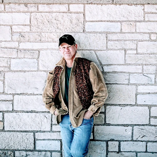 A man in casual attire, with a brown jacket and blue jeans, leans against a stone wall with his hands in his pockets, wearing a cap.