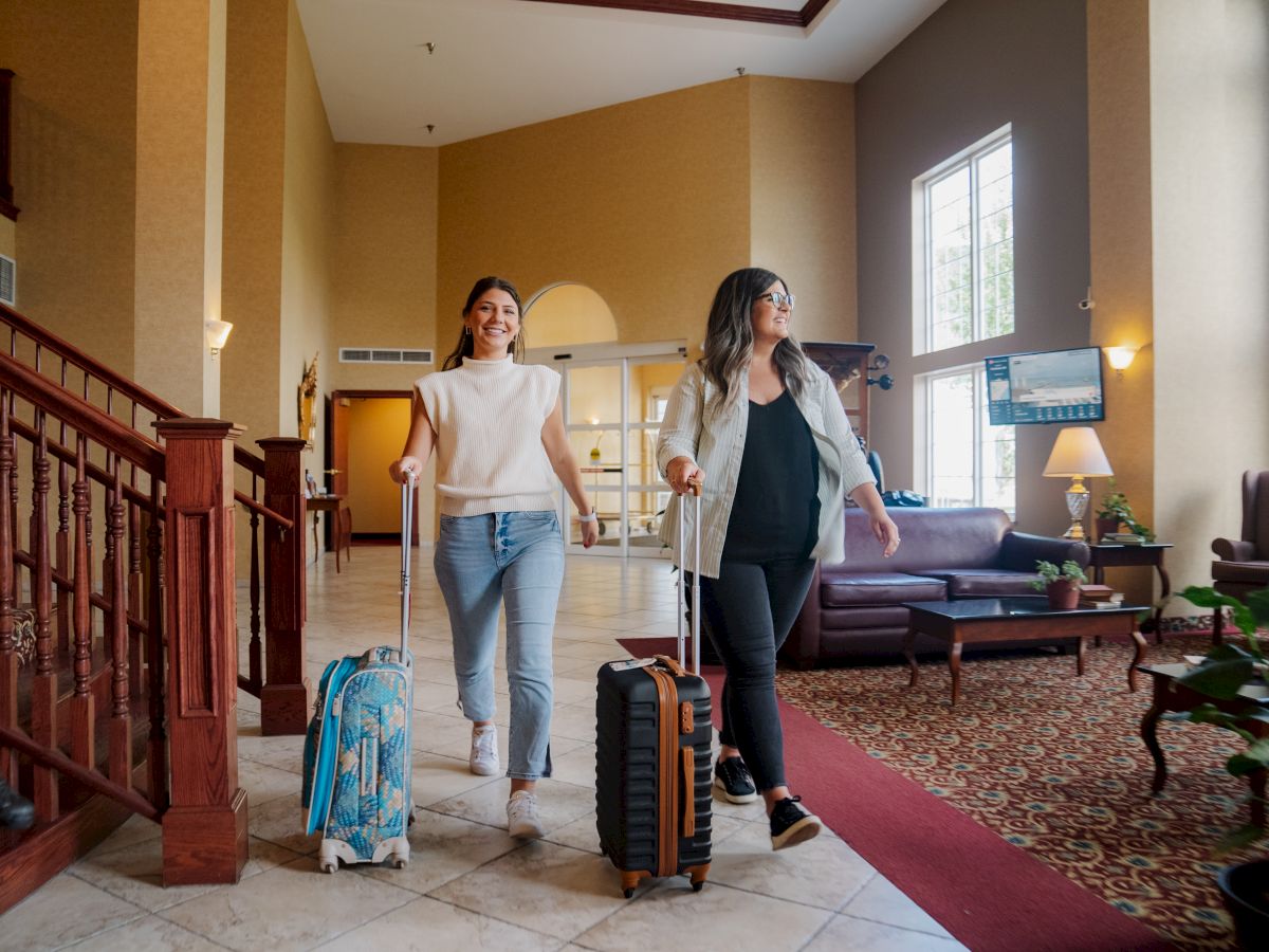 Two people with suitcases walk through a spacious, well-lit hotel lobby with a staircase, seating area, and large windows.