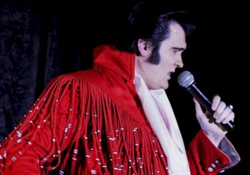 A person wearing a red, fringed jacket is holding a microphone and appears to be singing or performing against a dark background.