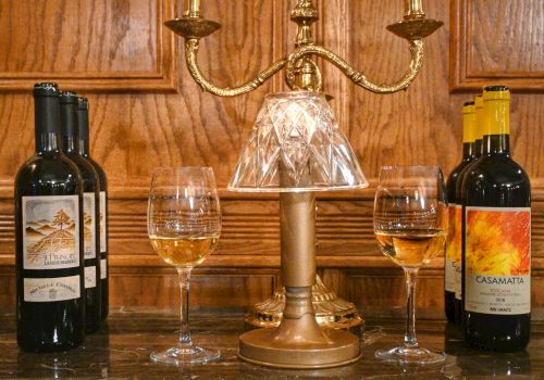 The image shows wine bottles with two glasses of wine on a table, flanking a gold lamp with crystal shade, set against a wooden background.