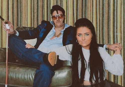 The image shows a man in a blue suit and sunglasses, and a woman with long dark hair, casually posed in front of a striped curtain.