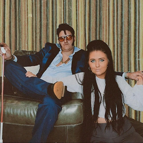The image shows a man in a blue suit and sunglasses, and a woman with long dark hair, casually posed in front of a striped curtain.