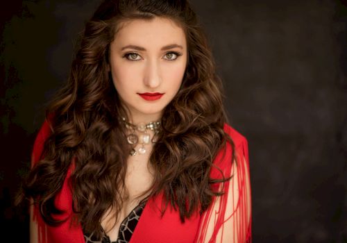 A woman with long, wavy hair wearing a red outfit and a necklace, against a dark background.