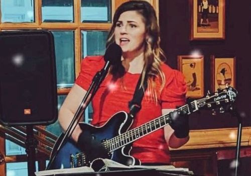 A woman in a red dress is playing an electric guitar and singing into a microphone at an indoor venue with framed pictures on the wall behind.
