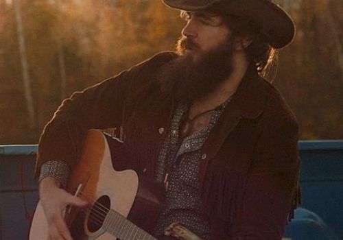 A person in a cowboy hat and jacket plays an acoustic guitar outdoors with a Coors Light can beside them in warm, evening light.