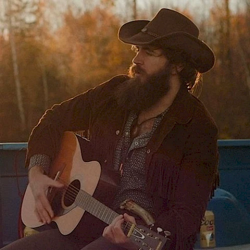 A person in a cowboy hat and jacket plays an acoustic guitar outdoors with a Coors Light can beside them in warm, evening light.