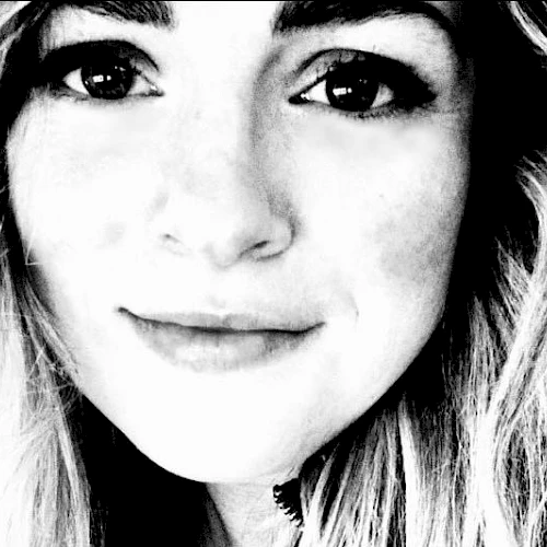 A black and white close-up photo of a person with long hair, light skin, and prominent eyes, looking directly at the camera and smiling subtly.