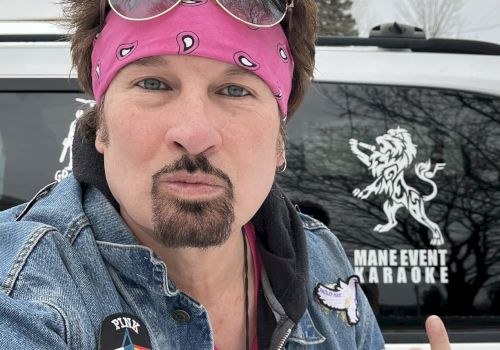 A person wearing a pink bandana and glasses poses in front of a vehicle with a "MAIN EVENT KARAOKE" logo and a lion decal, making a gesture.