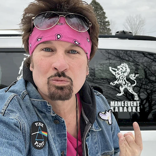 A person wearing a pink bandana and glasses poses in front of a vehicle with a "MAIN EVENT KARAOKE" logo and a lion decal, making a gesture.