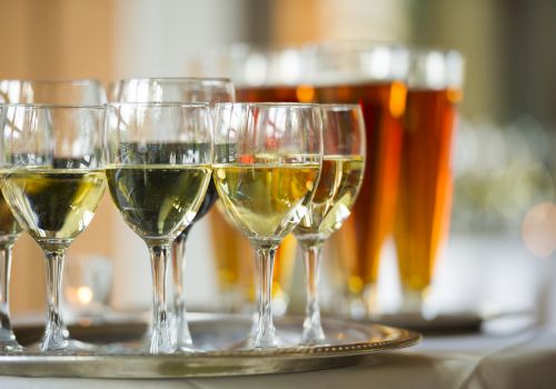 The image shows a tray with several glasses of white wine in the foreground and a row of beer glasses in the background on a table.