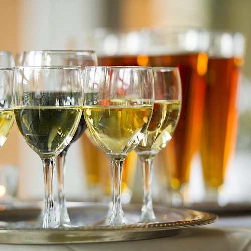 The image shows a tray with several glasses of white wine in the foreground and a row of beer glasses in the background on a table.