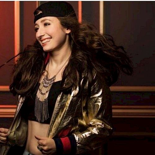 A person smiling, wearing a black cap backwards, a gold and black jacket, layered necklaces, and a crop top, standing against a wall with geometric accents.