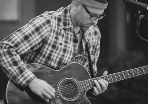 A person wearing glasses and a beanie is playing an acoustic guitar beside a microphone, dressed in a plaid shirt.