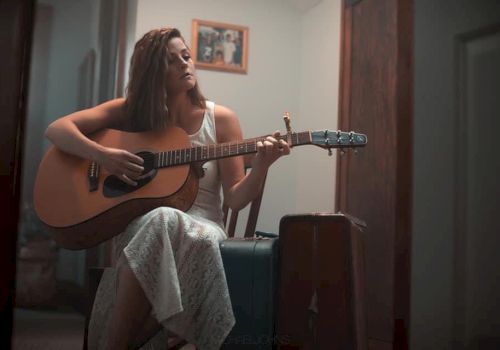 A person in a white dress is sitting on a chair and playing an acoustic guitar next to a suitcase, with a framed picture on the wall behind them.