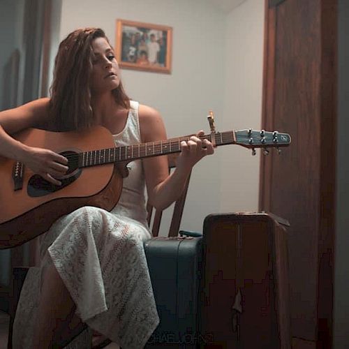 A person in a white dress is sitting on a chair and playing an acoustic guitar next to a suitcase, with a framed picture on the wall behind them.