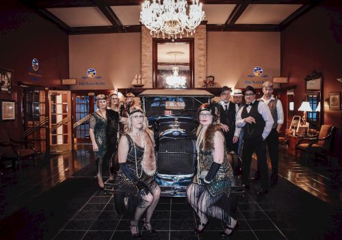 A group of people in 1920s attire pose in front of a vintage car inside a luxurious, chandelier-lit room, creating a glamorous setting.